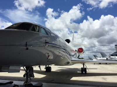 A large, sleek private jet is parked on a tarmac under a bright blue sky with scattered white clouds. Other smaller aircraft are visible in the background, and an airport control tower can be seen in the distance.