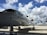 A large, sleek private jet is parked on a tarmac under a bright blue sky with scattered white clouds. Other smaller aircraft are visible in the background, and an airport control tower can be seen in the distance.