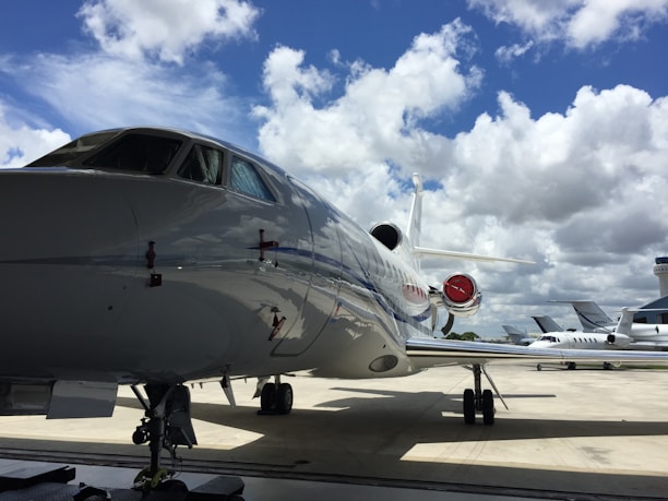 A large, sleek private jet is parked on a tarmac under a bright blue sky with scattered white clouds. Other smaller aircraft are visible in the background, and an airport control tower can be seen in the distance.