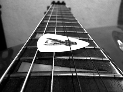 Black and white close-up image of a guitar fingerboard with a white guitar pick placed on the strings. The focus is on the strings and the pick, creating a depth effect along the guitar's neck.