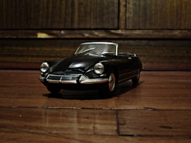 A detailed model of a classic black car is positioned on a wooden surface. The car has a shiny, reflective finish and distinctive round headlights. The background consists of a wooden panel, adding a rustic feel to the scene.