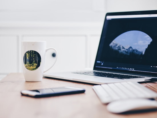 A wooden desk with a laptop displaying an image of mountains on its screen. A white mug with a circular forest design sits beside a smartphone and a keyboard. The setting conveys a minimalist workspace.