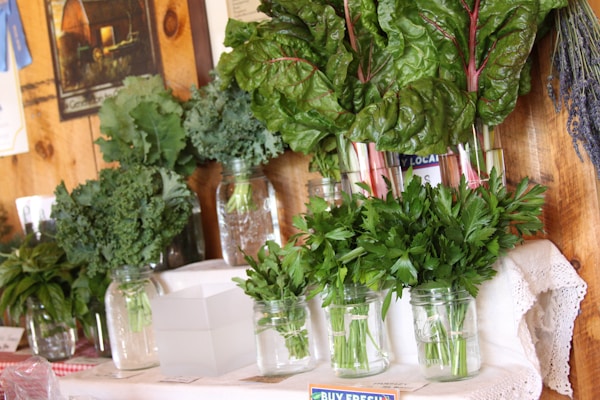 An assortment of fresh leafy greens is displayed in glass jars on a wooden shelf. The selection includes kale, Swiss chard, and parsley. A rustic setting with a picture on the wooden wall and a white lace tablecloth adds a natural and homely feel.
