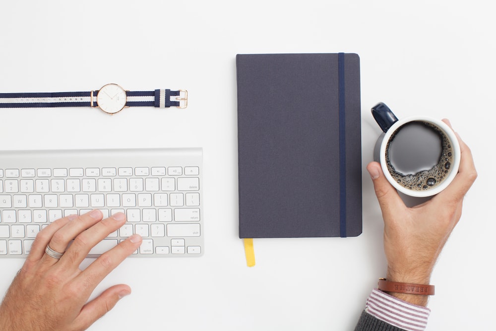 A man's hand holding a cup of coffee on a workspace with a notebook and a keyboard