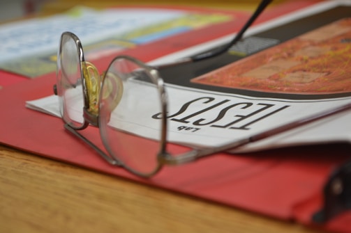 A pair of eyeglasses resting on top of a magazine with the headline 'Tests' visible. The magazine is placed on a red folder, and there is a blurred background with indistinguishable text and images. The scene suggests a casual setting, possibly in an office or study space.