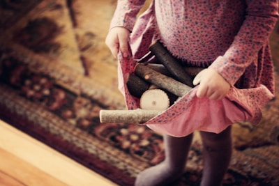 A child wearing a patterned dress is holding several pieces of firewood in the skirt of the dress. The background features a patterned rug with warm tones.