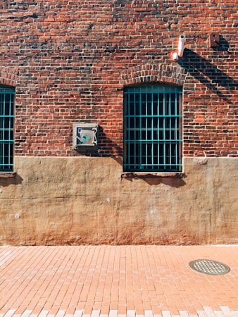 A brick wall with two windows that have teal metal bars. The wall is divided into a red brick upper section and a beige concrete lower section. A small utility box is mounted on the wall between the windows, and a security camera is positioned above, casting a shadow.
