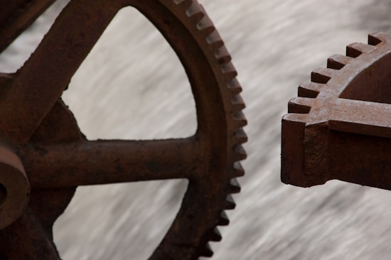 Two large, rusty iron gears are positioned closely together, showing detailed teeth that mesh with each other. The background is blurred, focusing attention on the mechanical components.