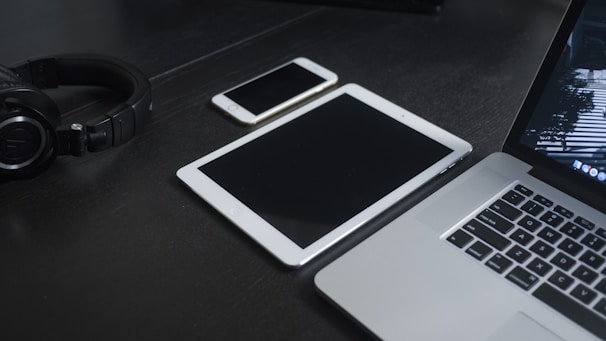A dark surface holds various electronic devices including a pair of black headphones, a smartphone, a tablet, and a laptop. The arrangement is neat and orderly, with the devices laid out flat.