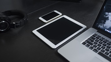 A dark surface holds various electronic devices including a pair of black headphones, a smartphone, a tablet, and a laptop. The arrangement is neat and orderly, with the devices laid out flat.