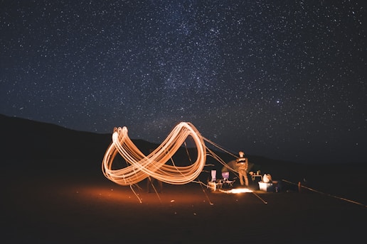 Under a clear starry sky, a campsite is illuminated by swirling patterns of light created by long-exposure photography. A person stands next to camping chairs and gear, with the night sky providing a vast, cosmic backdrop.