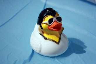 A rubber duck dressed in a stylized black wig and white sunglasses, resembling a rock and roll icon. The duck is positioned on a light blue surface, suggesting a playful theme.