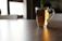 A clear glass cup filled with a warm amber-colored liquid is placed on a wooden table. The background is softly blurred, showing parts of a chair and window, suggesting a peaceful indoor setting.