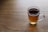 A clear glass mug filled with dark amber tea placed on a wooden surface. The tea has a warm, inviting appearance with a few small bubbles visible on the surface. Soft natural light casts a gentle shadow on the table.