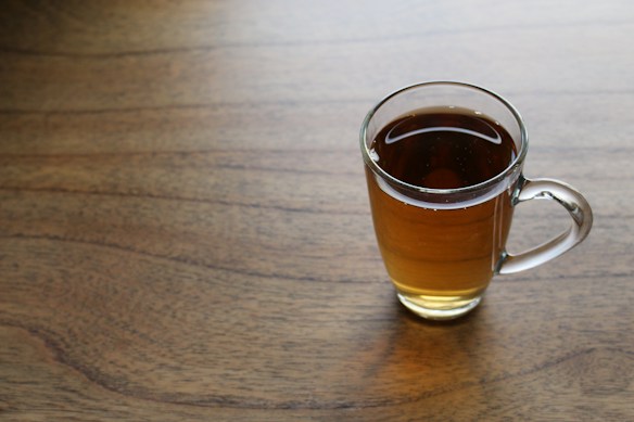 A clear glass mug filled with dark amber tea placed on a wooden surface. The tea has a warm, inviting appearance with a few small bubbles visible on the surface. Soft natural light casts a gentle shadow on the table.