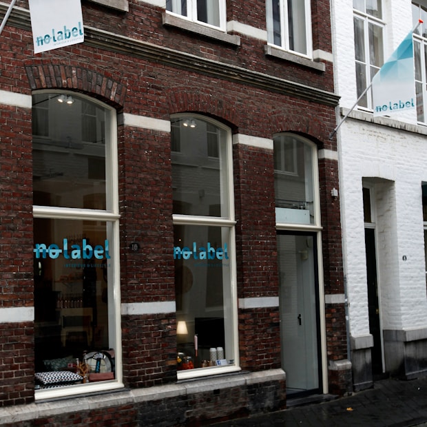 The storefront of a shop with large windows in a brick building, featuring the words 'no label' in blue on the glass. Displayed inside are various products, including bags and decorative items, with a lamp also visible. The facade has a combination of brick and white sections, and there is a small sign protruding from the wall.
