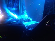 A DJ is performing in a dimly lit setting, surrounded by neon blue lights. The image captures turntables, a laptop with a glowing logo, and the DJ's hands in motion, suggesting an energetic atmosphere.