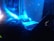 A DJ is performing in a dimly lit setting, surrounded by neon blue lights. The image captures turntables, a laptop with a glowing logo, and the DJ's hands in motion, suggesting an energetic atmosphere.