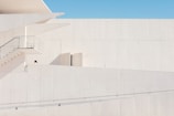 A minimalist architectural structure exhibits clean lines and a modern design. The image is dominated by pale, light-colored walls with a small section of a staircase and glass railing visible. One person, partially seen, appears on the first level of the building.