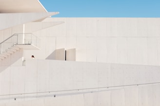 A minimalist architectural structure exhibits clean lines and a modern design. The image is dominated by pale, light-colored walls with a small section of a staircase and glass railing visible. One person, partially seen, appears on the first level of the building.