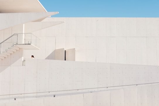 A minimalist architectural structure exhibits clean lines and a modern design. The image is dominated by pale, light-colored walls with a small section of a staircase and glass railing visible. One person, partially seen, appears on the first level of the building.