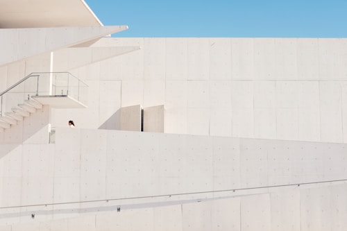 A minimalist architectural structure exhibits clean lines and a modern design. The image is dominated by pale, light-colored walls with a small section of a staircase and glass railing visible. One person, partially seen, appears on the first level of the building.