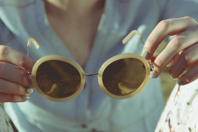 Hands holding a pair of round, vintage-style sunglasses with tinted lenses. The background is softly blurred, and the lighting gives a warm, vintage feel. The person is wearing a light-colored shirt.