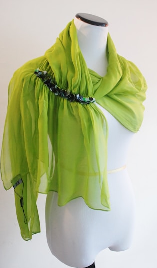 A vibrant lime green sheer scarf with decorative embellishments is elegantly draped over a white dress form. The scarf features pleating and a row of dark sequins or beads near the top.