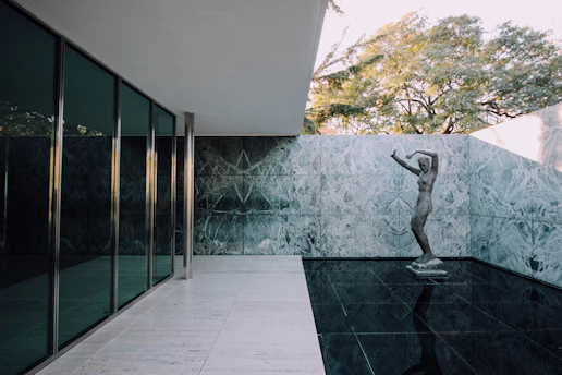 A modern architectural setting featuring a minimalist design with large glass panels on the left, a marble wall in the center, and a reflective dark floor creating a sense of openness and lightness. A classical statue stands at the intersection of the floor and wall, surrounded by the tranquil environment. In the background, tall trees can be seen over the wall, adding a touch of nature to the industrial setting.