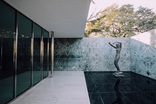 A modern architectural setting featuring a minimalist design with large glass panels on the left, a marble wall in the center, and a reflective dark floor creating a sense of openness and lightness. A classical statue stands at the intersection of the floor and wall, surrounded by the tranquil environment. In the background, tall trees can be seen over the wall, adding a touch of nature to the industrial setting.