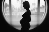 A silhouette of a pregnant woman standing in front of a large window with vertical blinds. The contrast between her figure and the bright outdoor light creates a striking image.