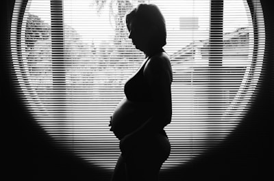 A silhouette of a pregnant woman standing in front of a large window with vertical blinds. The contrast between her figure and the bright outdoor light creates a striking image.