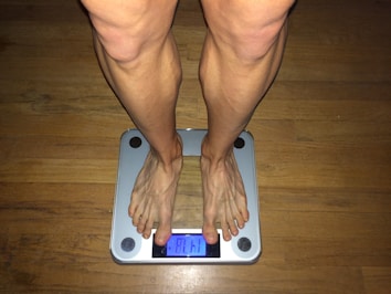 A person's legs and feet standing on a digital weighing scale. The display reads '81.1'. The floor underneath is wooden, and the scale has a metallic frame with four circular rubber accents at each corner.