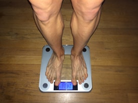 A person's legs and feet standing on a digital weighing scale. The display reads '81.1'. The floor underneath is wooden, and the scale has a metallic frame with four circular rubber accents at each corner.