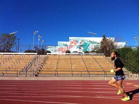 A runner with a beard is jogging on a red track next to an empty stadium. The sky is clear blue, and there are some trees and floodlights in the background. A large mural depicting athletes is visible behind the stands.