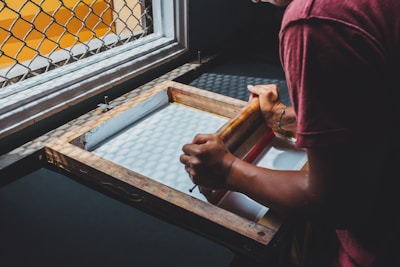 A person engaged in the process of screen printing, using a wooden frame and a mesh screen to apply ink onto a surface. The scene is lit by natural light coming through a window with a patterned grill, casting shadows on the workspace.
