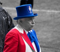 An elderly man is dressed in a vibrant outfit that prominently features the colors red, white, and blue. He wears a striking blue top hat with an emblem featuring a rooster and the letters 'FFR'. The man stands outdoors, possibly at a public or sports event.