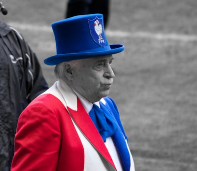 An elderly man is dressed in a vibrant outfit that prominently features the colors red, white, and blue. He wears a striking blue top hat with an emblem featuring a rooster and the letters 'FFR'. The man stands outdoors, possibly at a public or sports event.