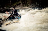 A group of people is engaged in whitewater rafting, navigating through turbulent rapids. They are wearing helmets and life jackets for safety, with paddles in hand. The white water splashes energetically around them, adding to the sense of movement and adventure.
