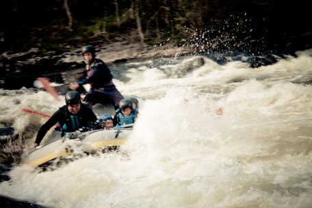 A group of people is engaged in whitewater rafting, navigating through turbulent rapids. They are wearing helmets and life jackets for safety, with paddles in hand. The white water splashes energetically around them, adding to the sense of movement and adventure.
