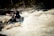 A group of people is engaged in whitewater rafting, navigating through turbulent rapids. They are wearing helmets and life jackets for safety, with paddles in hand. The white water splashes energetically around them, adding to the sense of movement and adventure.