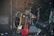 A collection of musical instruments including two electric guitars resting against a drum kit. The drums have metallic finishes and are surrounded by stands and microphones. A large stage light is positioned at the front, with an amplifier and water bottle visible in the background.