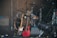 A collection of musical instruments including two electric guitars resting against a drum kit. The drums have metallic finishes and are surrounded by stands and microphones. A large stage light is positioned at the front, with an amplifier and water bottle visible in the background.