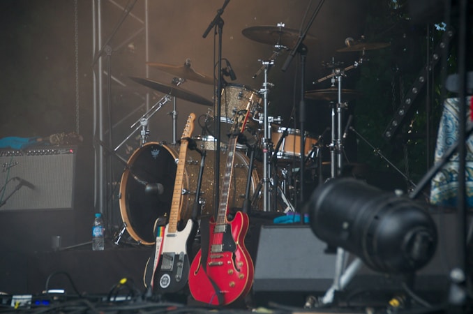 A collection of musical instruments including two electric guitars resting against a drum kit. The drums have metallic finishes and are surrounded by stands and microphones. A large stage light is positioned at the front, with an amplifier and water bottle visible in the background.