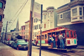 A vintage-style scene captures a classic San Francisco cable car full of passengers traveling up a steep street. Historic buildings line the road, with the Transamerica Pyramid visible in the background. The street sign and parked cars add a touch of urban life, while the scene has a nostalgic, warm tone.