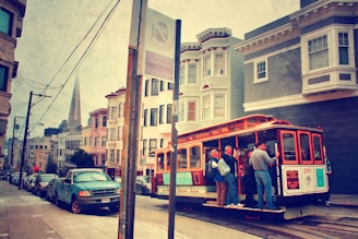 A vintage-style scene captures a classic San Francisco cable car full of passengers traveling up a steep street. Historic buildings line the road, with the Transamerica Pyramid visible in the background. The street sign and parked cars add a touch of urban life, while the scene has a nostalgic, warm tone.