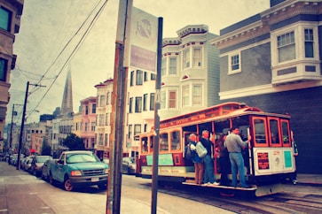 A vintage-style scene captures a classic San Francisco cable car full of passengers traveling up a steep street. Historic buildings line the road, with the Transamerica Pyramid visible in the background. The street sign and parked cars add a touch of urban life, while the scene has a nostalgic, warm tone.