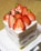 A square slice of layered cake topped with fresh strawberries and small green pistachios. The layers alternate between a light sponge and creamy filling, with visible pink strawberry layers.