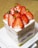 A square slice of layered cake topped with fresh strawberries and small green pistachios. The layers alternate between a light sponge and creamy filling, with visible pink strawberry layers.