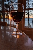 A wine glass filled with red wine is positioned on a table indoors, with the backdrop of a scenic sunset over a body of water. Sunlight streams through the trees and the window, casting reflections and creating a warm ambiance. The surrounding area is tranquil, with the silhouette of trees against the colorful sky.